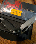 A Microtech folding knife with a textured black handle and a satin silver blade, presented in its black foam-lined box with the branded outer packaging and a yellow sharp edge warning sticker.