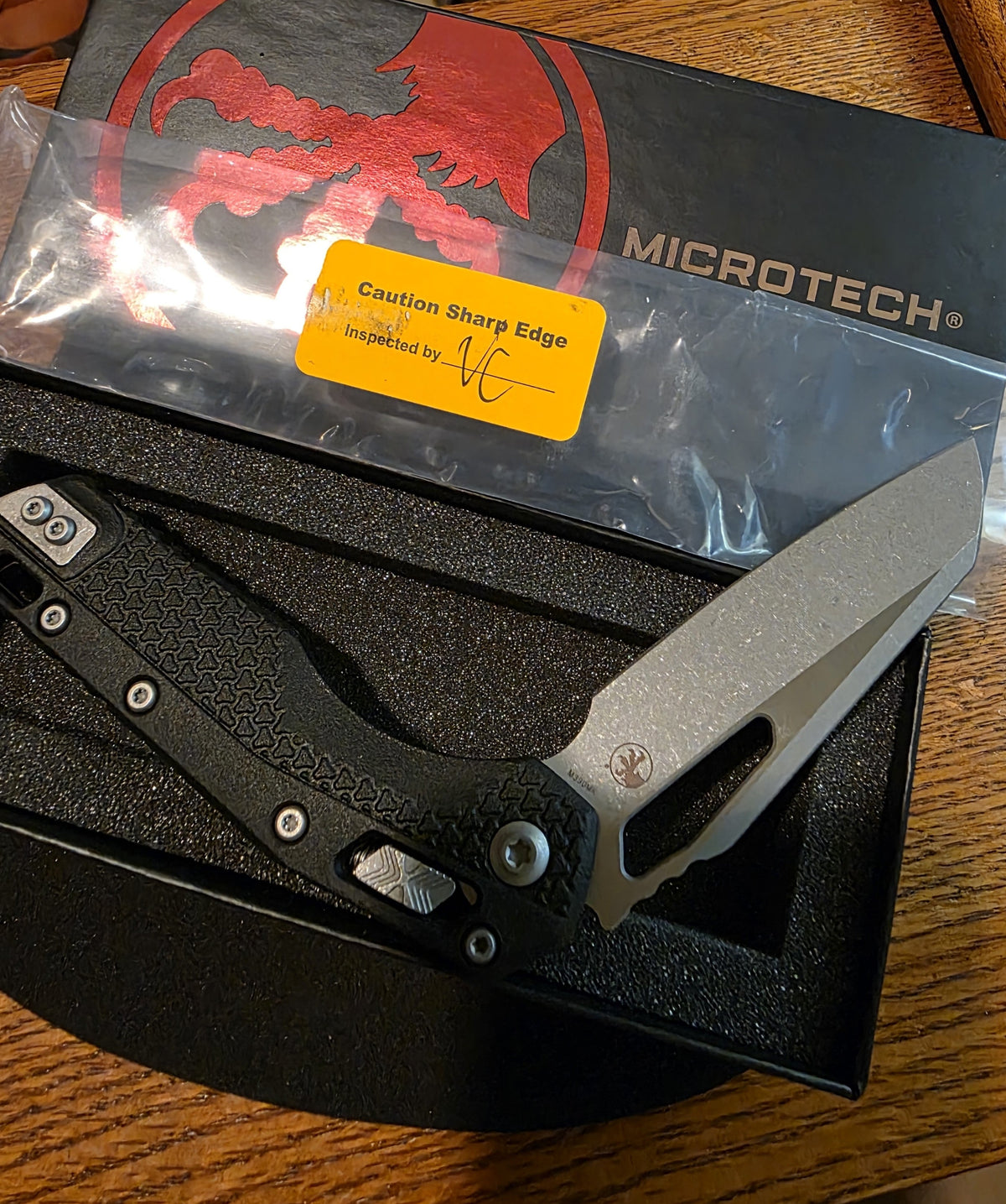A Microtech folding knife with a textured black handle and a satin silver blade, presented in its black foam-lined box with the branded outer packaging and a yellow sharp edge warning sticker.