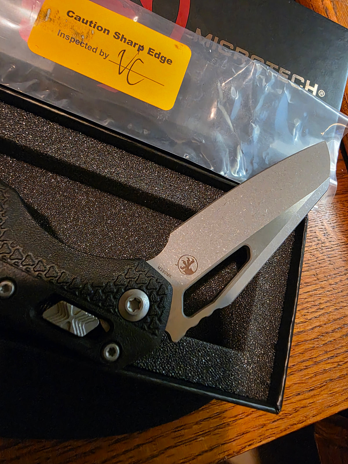 A folding pocket knife with a textured black handle and a matte silver blade, presented in its black foam-lined box. A yellow 'Caution Sharp Edge' sticker is visible on the plastic packaging, and the brand name 'MIDDBYTECH' is printed on the box lid.