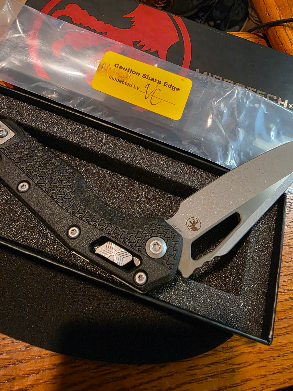 A folding pocket knife with a textured black handle and a silver blade, displayed inside its black presentation box. A yellow sticker on the plastic wrap reads 'Caution Sharp Edge' and is signed by Mikaele.