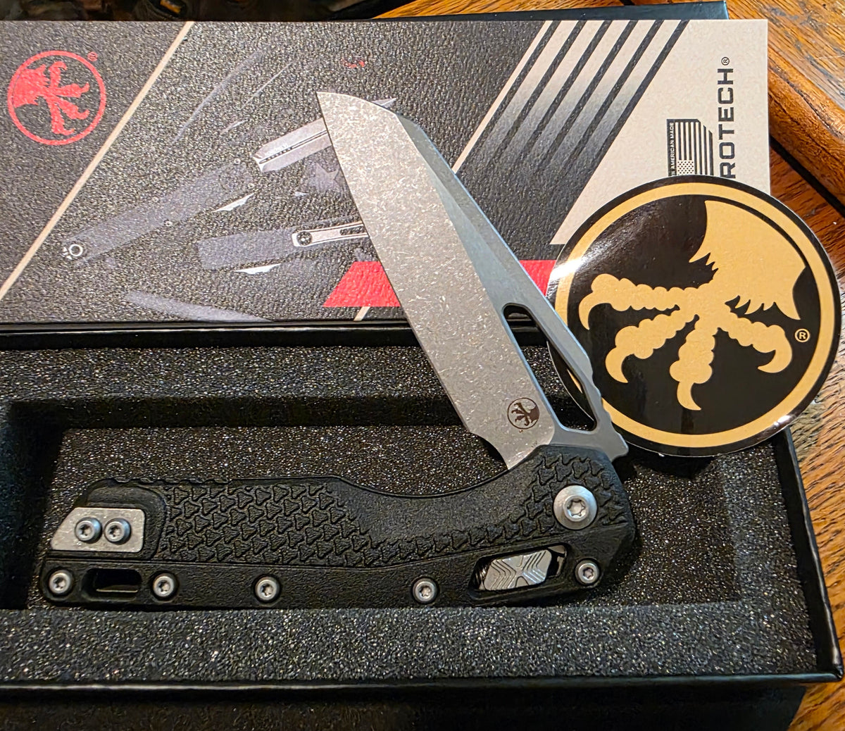 A black and silver folding pocket knife with a textured handle and a glittery silver blade, displayed in its open black presentation box. The box lid features the ROTECH brand logo, and a round black-and-gold ROTECH sticker is placed next to the knife on a wooden table.