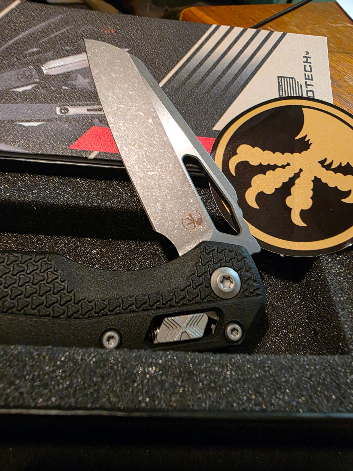 A folding pocket knife with a textured black handle and a satin-finish silver blade, displayed inside a black foam presentation box. A circular black and gold medallion with a stylized bird logo is visible next to the knife, along with branded packaging in the background.