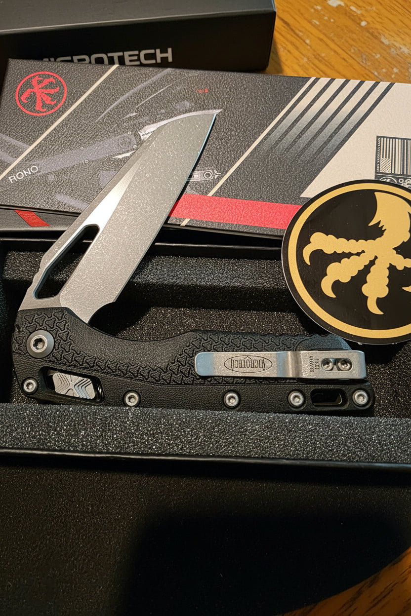 A folding pocket knife with a textured black handle and a matte gray blade, displayed inside a black presentation box alongside a circular black and gold emblem.