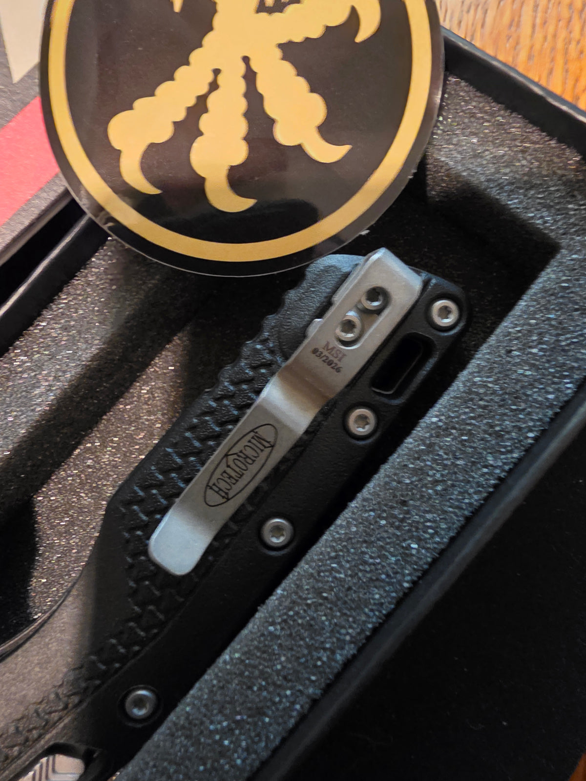 A close-up of a black folding knife with a silver metal clip, resting inside a black foam-lined presentation box. A round black and gold medallion with a claw-like emblem is visible at the top.