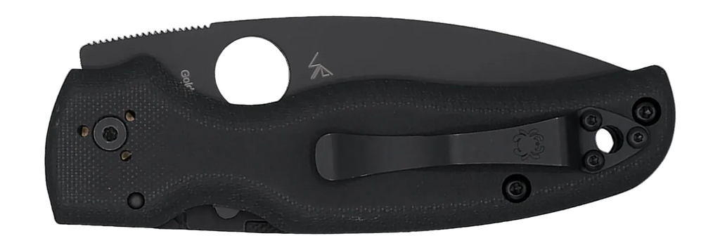 A black folding knife shown in its sheath against a plain white background. The knife features a textured black handle, a black pocket clip, and a partially serrated back edge.
