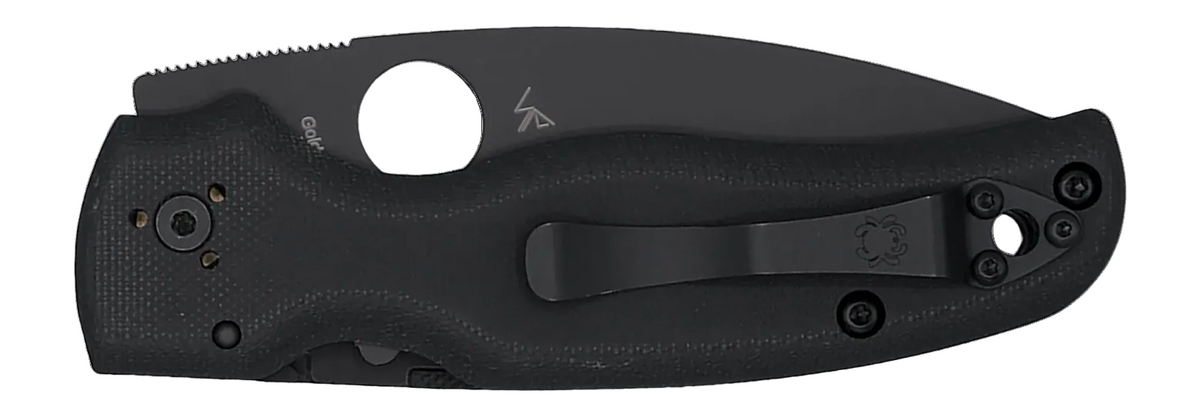 A black folding knife shown in its sheath against a plain white background. The knife features a textured black handle, a black pocket clip, and a partially serrated back edge.