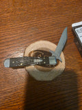A folding pocket knife with a silver blade and a dark patterned handle, resting on a small round wooden coaster on a wooden table.