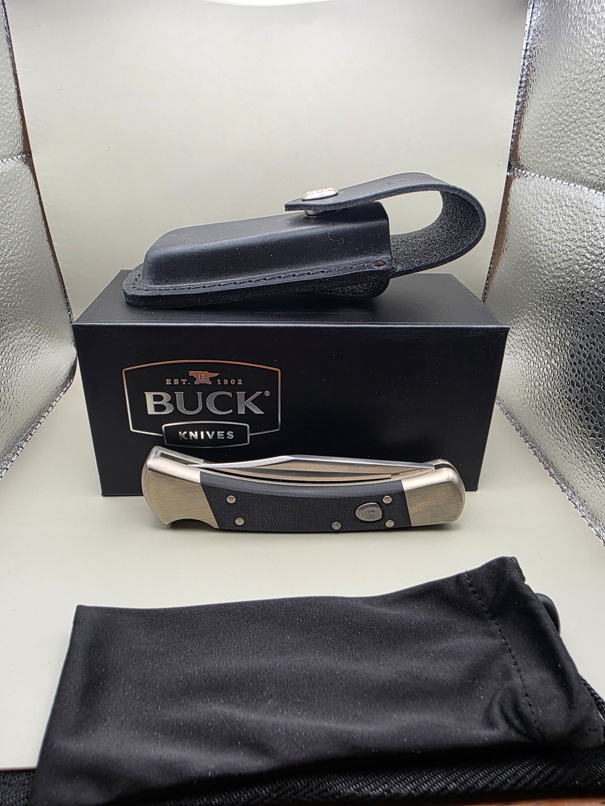 A folding pocket knife with a black handle and silver-toned metal bolster, displayed next to its black presentation box, a black leather sheath, and a black fabric pouch on a light surface.