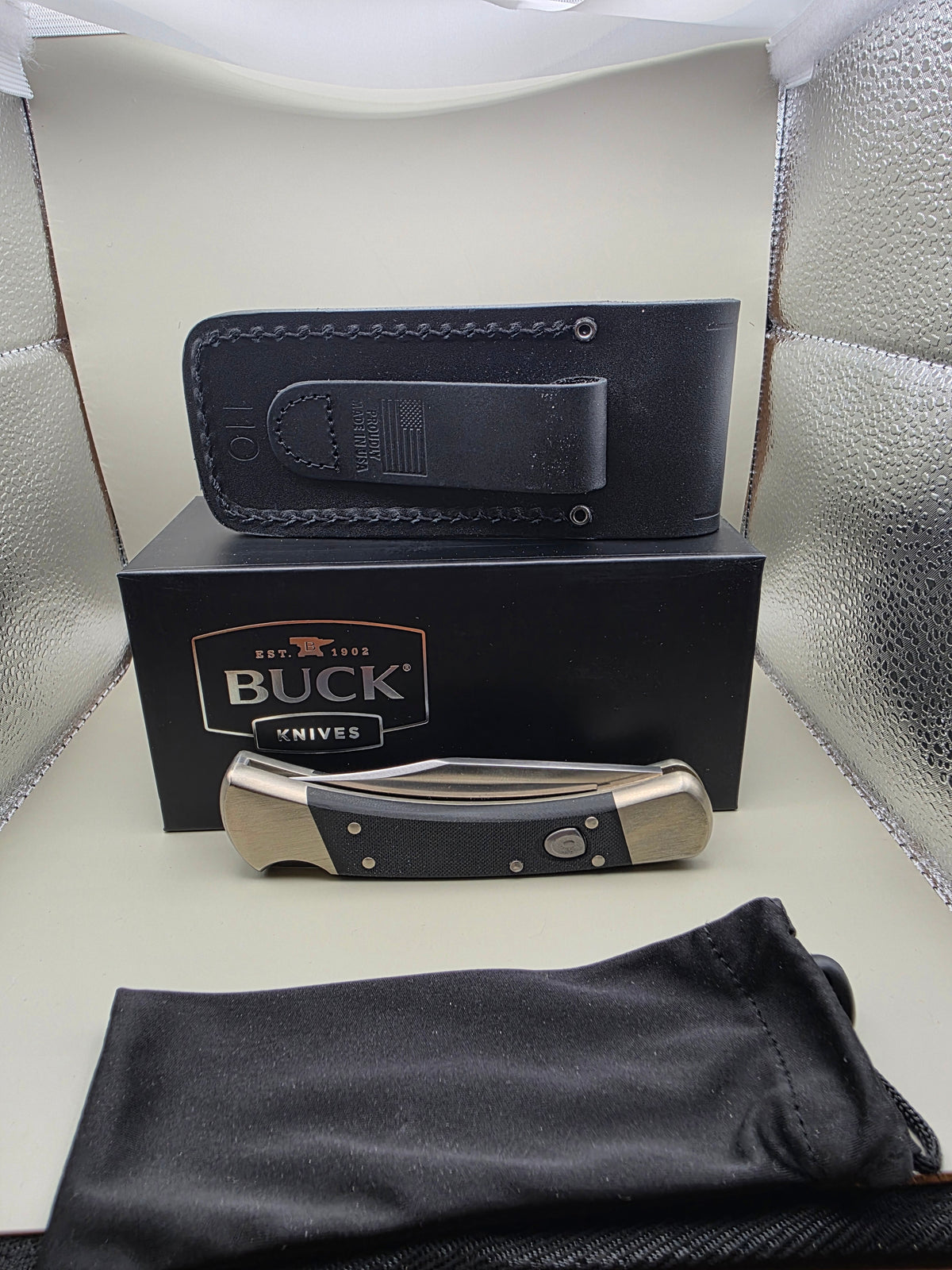 A folding pocket knife with a black handle and silver metal blade, displayed next to its black presentation box, a black leather sheath, and a black drawstring pouch on a light-colored surface.