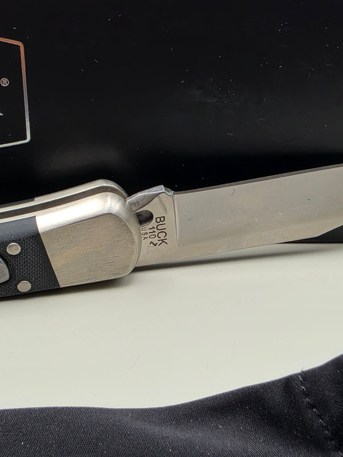 A close-up of a folding pocket knife with a silver blade and handle, featuring the 'BUCK' brand name engraved on the blade, resting against a black and white background.