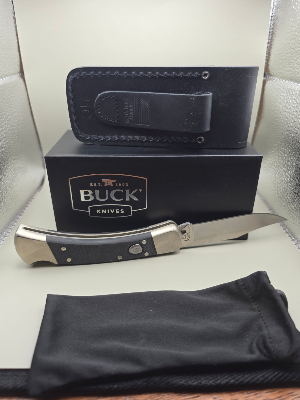 A Buck Knives folding pocket knife with a black handle and silver blade, displayed next to its black presentation box, a black leather sheath, and a black fabric pouch on a light-colored surface.