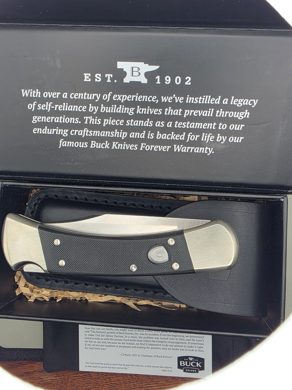 A folding pocket knife with a black textured handle and silver metal blade, displayed inside an open black presentation box with printed company text and a warranty card. The Buck Knives logo is visible on the card.