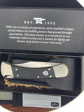 A folding pocket knife with a black textured handle and silver metal blade, displayed inside an open black presentation box with printed company text and a warranty card. The Buck Knives logo is visible on the card.