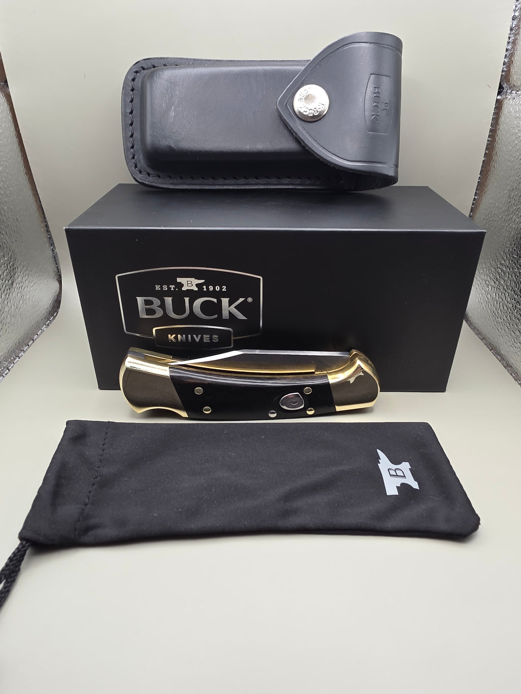 A folding pocket knife with black and gold scales is displayed next to its black presentation box, a black leather sheath, and a black drawstring pouch with a white Buck logo, all arranged on a light gray surface.