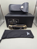A folding pocket knife with black and gold scales is displayed next to its black presentation box, a black leather sheath, and a black drawstring pouch with a white Buck logo, all arranged on a light gray surface.