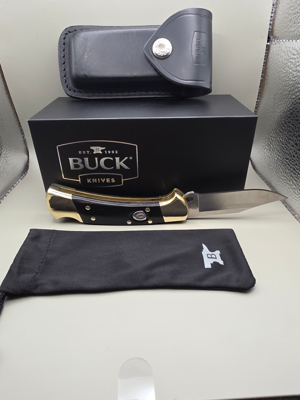 A Buck Knives folding knife with a black handle and brass bolster, displayed next to its black presentation box, a black leather sheath, and a black fabric pouch on a light-colored surface.