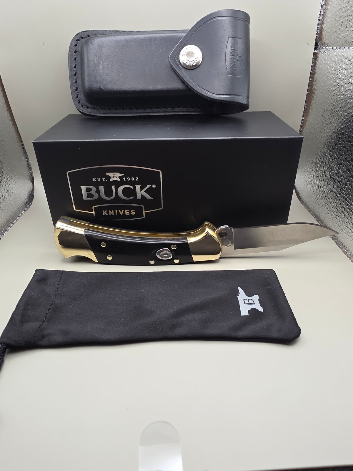A Buck Knives folding knife with a black handle and brass bolster, displayed next to its black presentation box, a black leather sheath, and a black fabric pouch on a light-colored surface.
