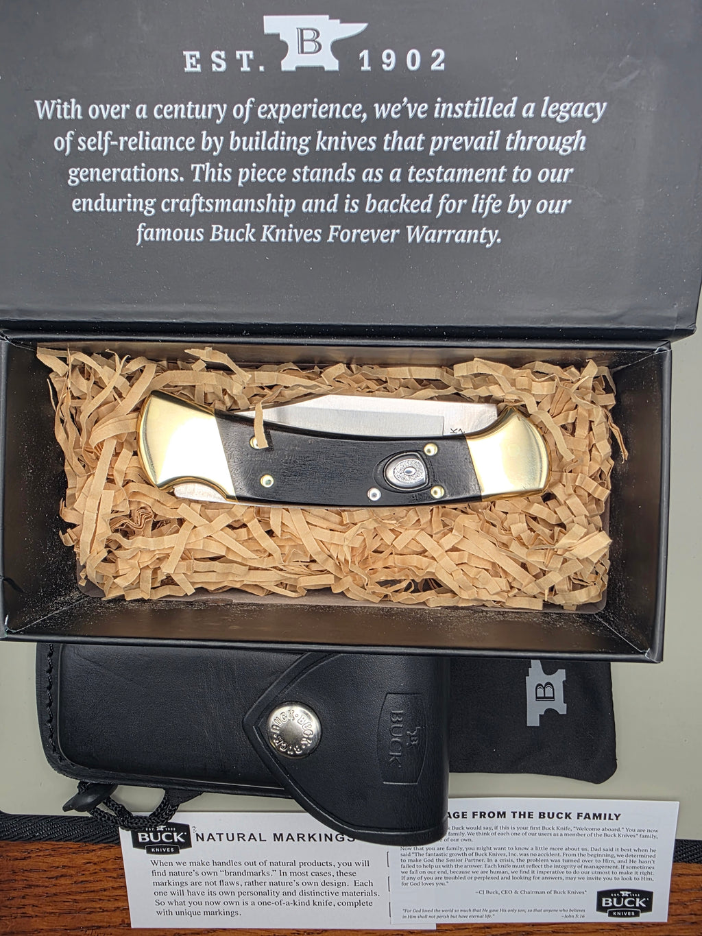 A folding pocket knife with black handles and brass-toned end caps is displayed inside an open presentation box filled with shredded paper. A black leather knife sheath, warranty card, and branded box are arranged around it on a tabletop.
