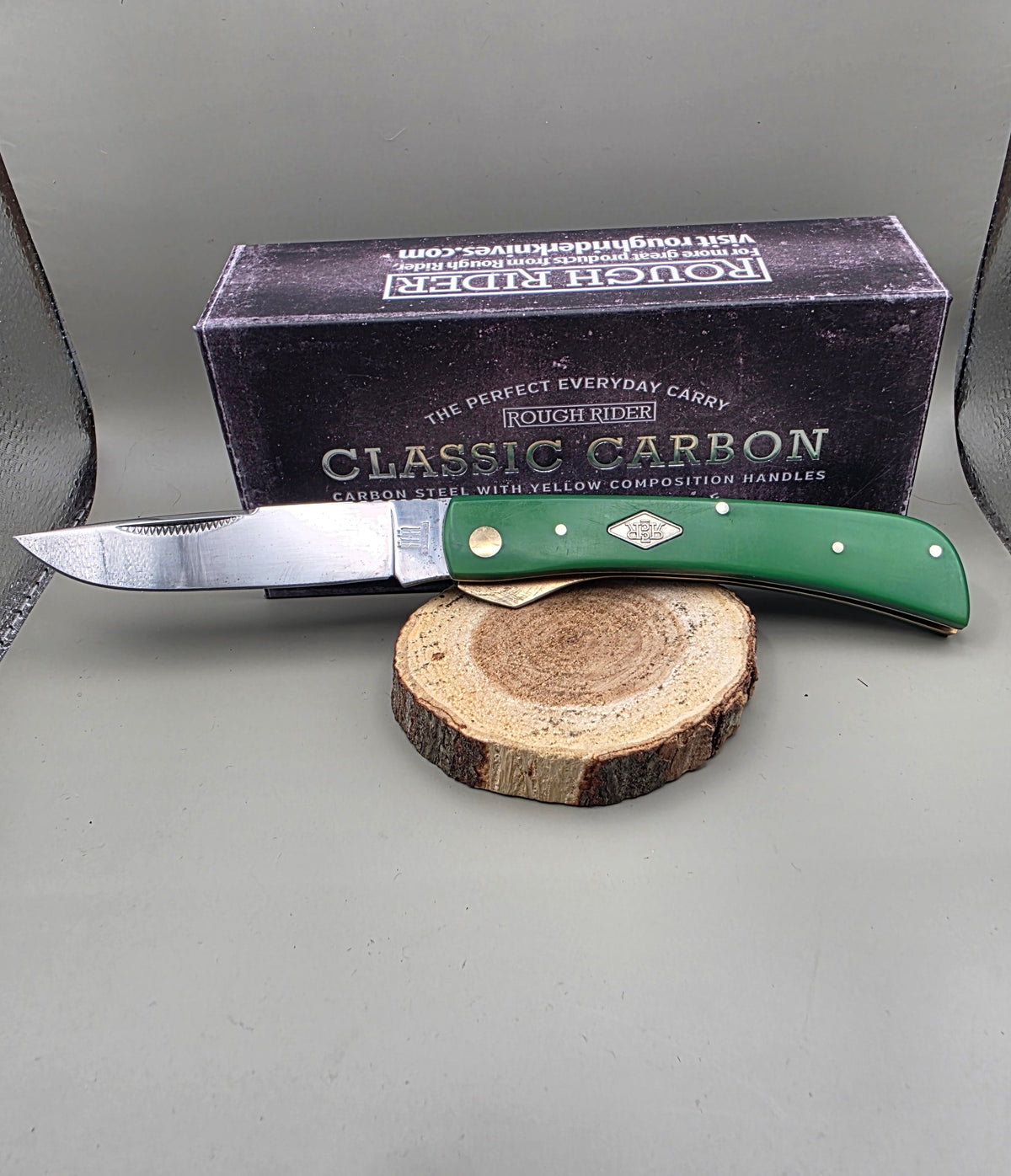 A pocket knife with a green handle and silver blade rests on a round wood slice in front of its black packaging box. The box is labeled 'BOSTON SHIP' and 'CLASSIC CARBON'.