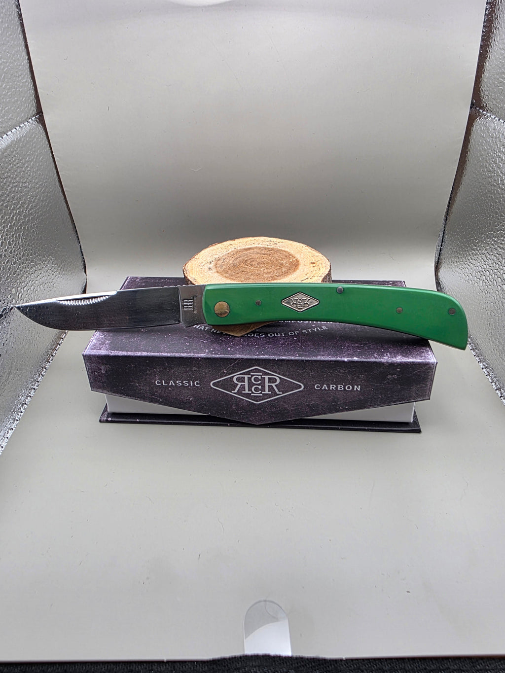 A folding pocket knife with a green handle and silver blade rests on top of a dark purple A&R Classic Carbon box, with a small wooden stand behind it, set inside a silver textured display case.