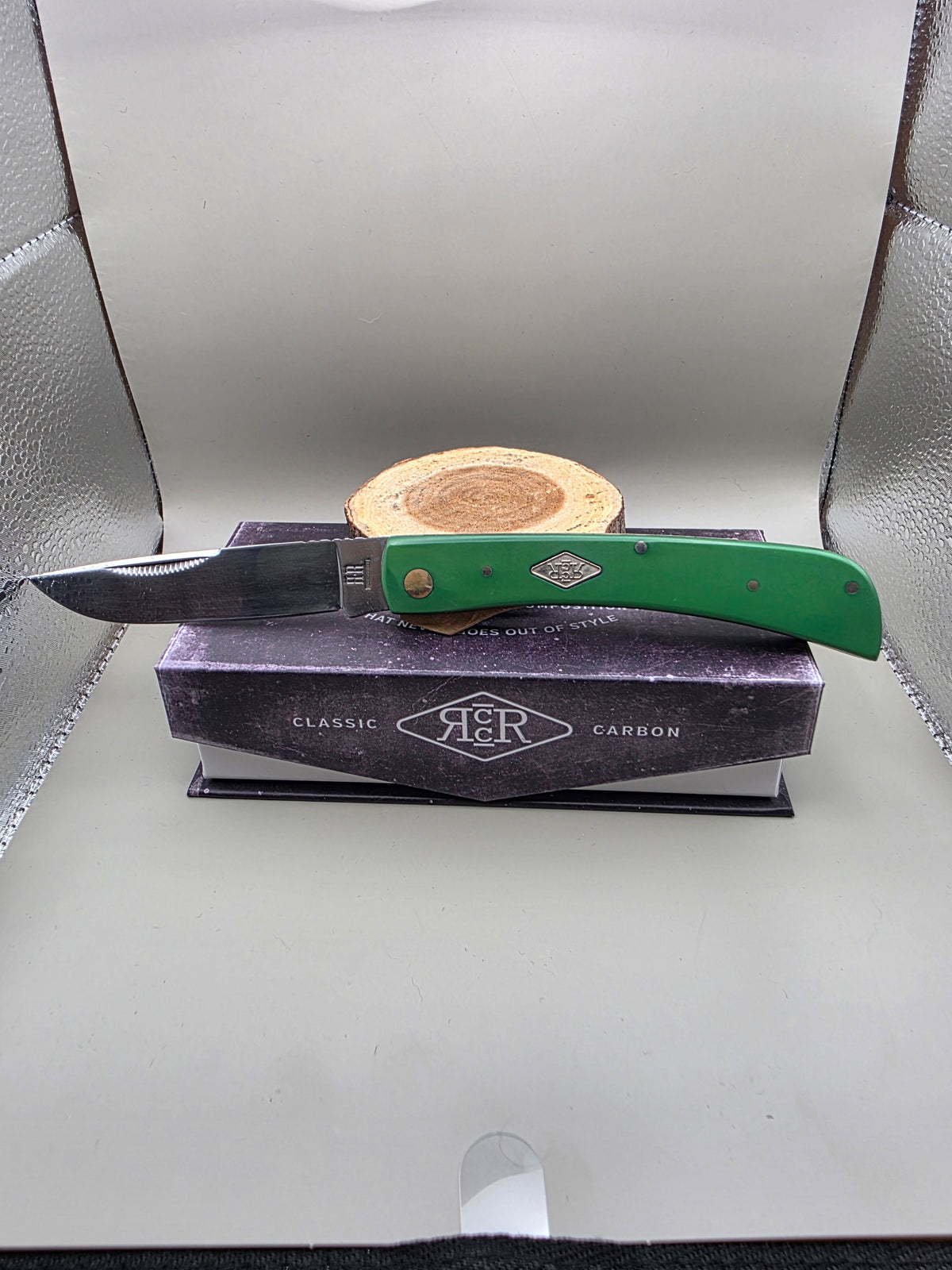 A folding pocket knife with a green handle and silver blade rests on top of a dark purple A&R Classic Carbon box, with a small wooden stand behind it, set inside a silver textured display case.