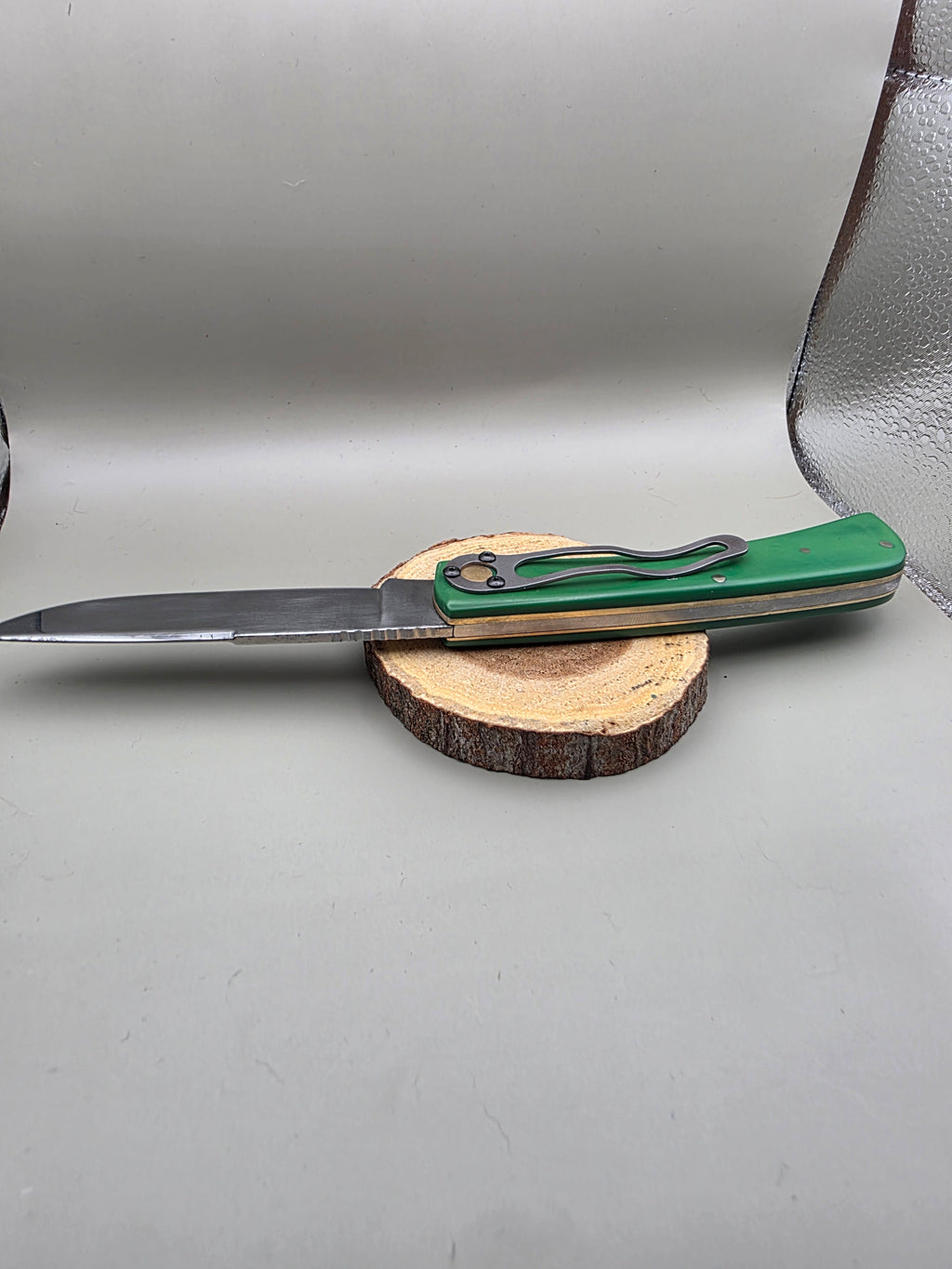 A folding pocket knife with a green handle and silver blade, resting on a round wood slice stand against a light gray background.