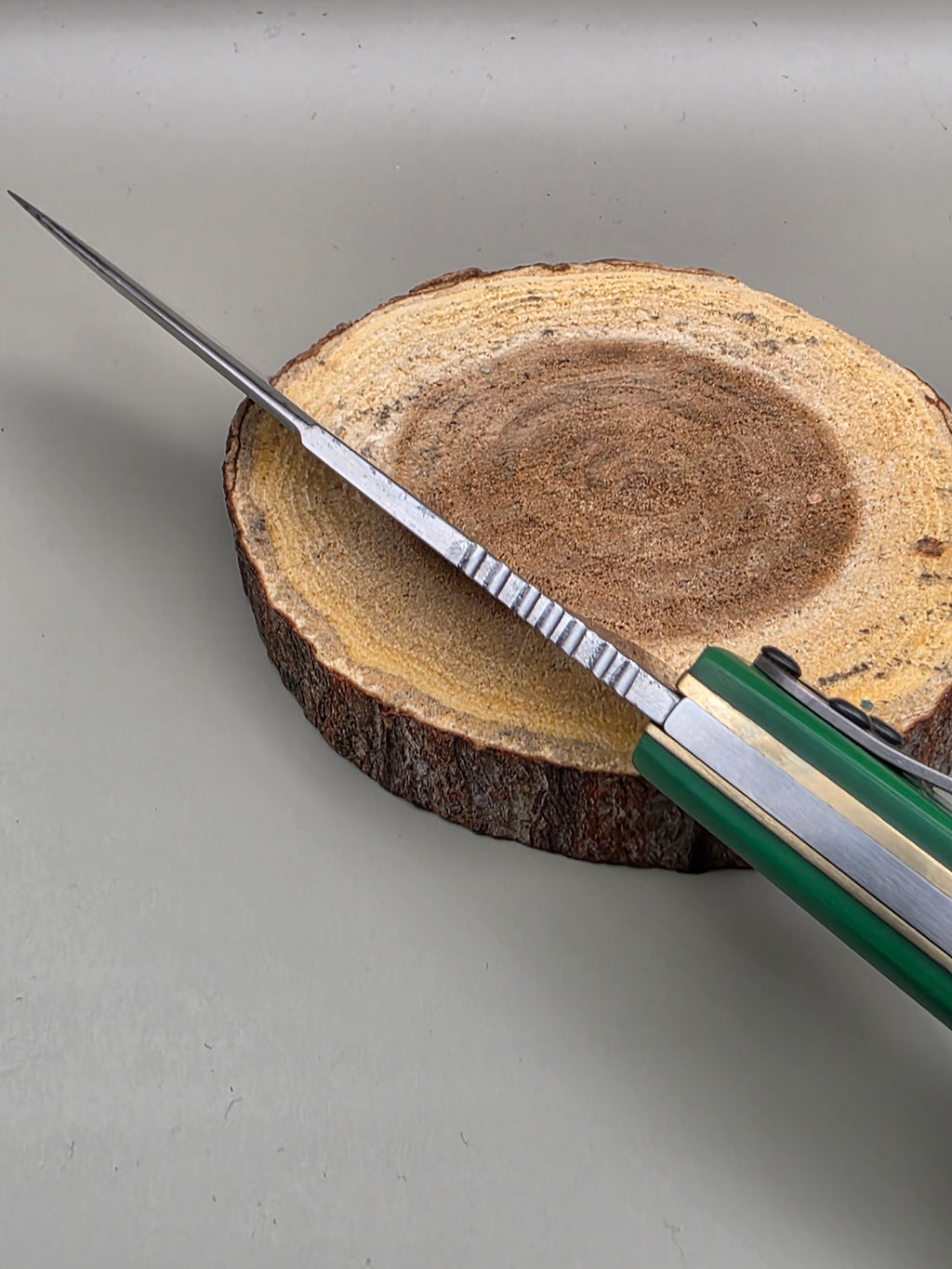 A folding pocket knife with a green and silver handle is partially opened, resting diagonally on a round wood slice prop against a neutral gray background.
