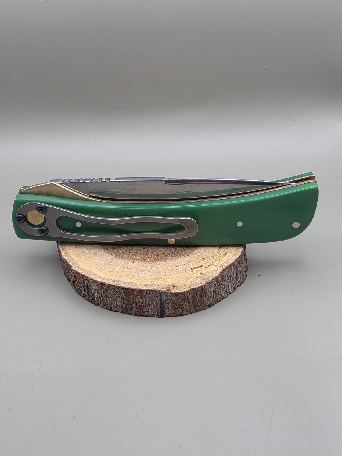 A folding pocket knife with a green handle and a dark blade, displayed on a round wood slice against a neutral gray background.