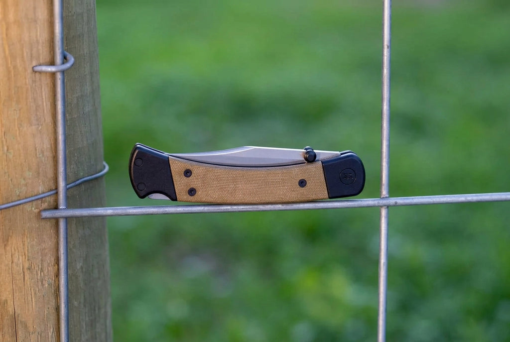 A folding pocket knife with a satin finish steel blade and a textured tan handle with black accents, shown resting against a wire fence on a wooden post with a blurred green grass background. 