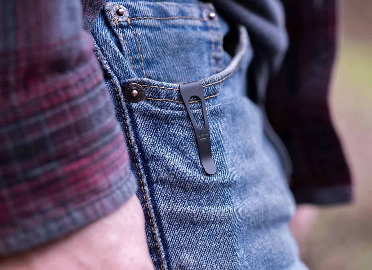 Close-up of a person wearing blue denim jeans with a dark metal knife sheath in the pocket, along with a plaid shirt and blurred outdoor background. 