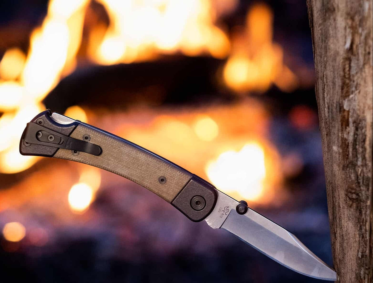 A folding pocket knife with a textured tan handle and a silver blade is shown partially inserted into a tree trunk, with a blurred campfire in the background. 