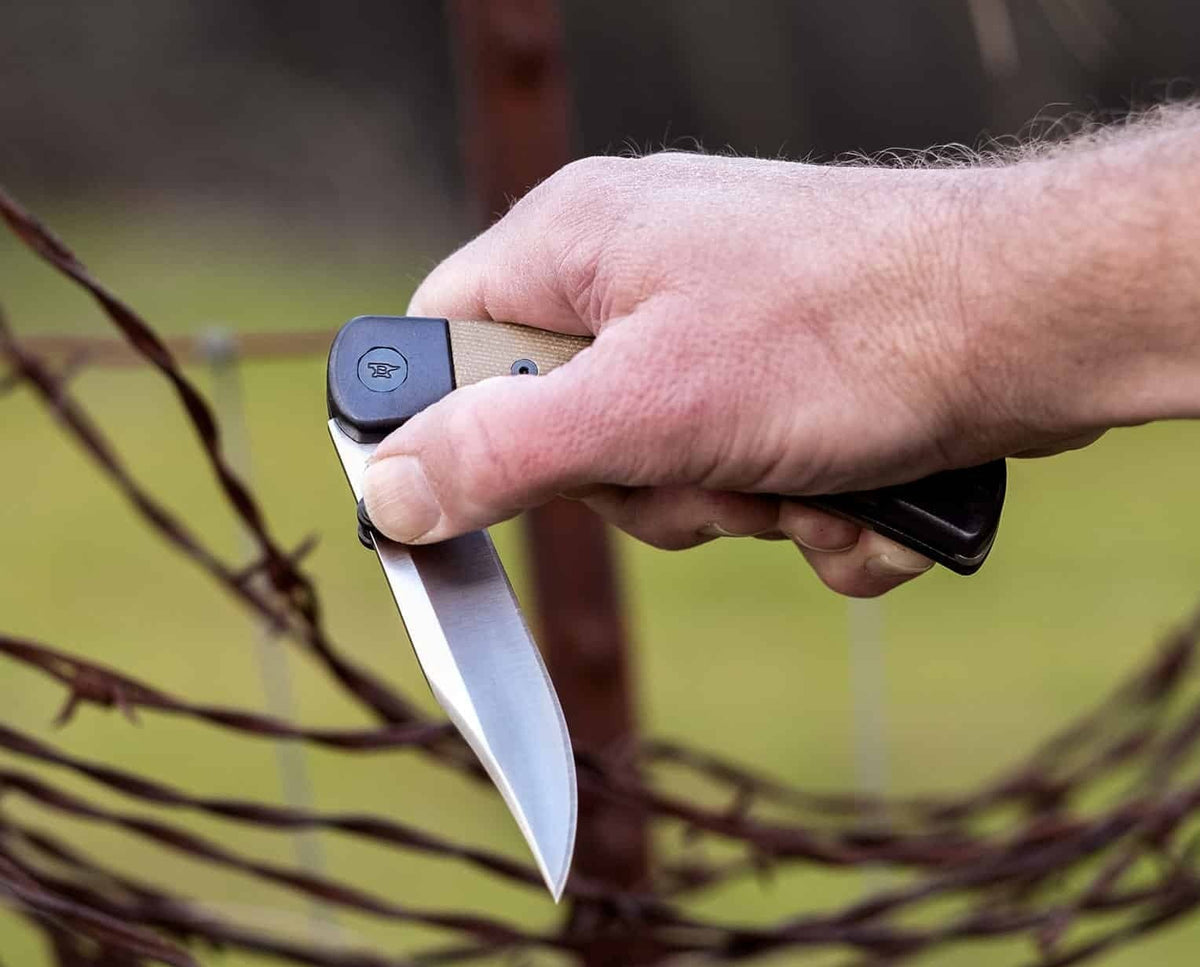 A close-up of a hand holding a folding pocket knife with a silver blade and tan handle in front of a blurred barbed wire fence.
