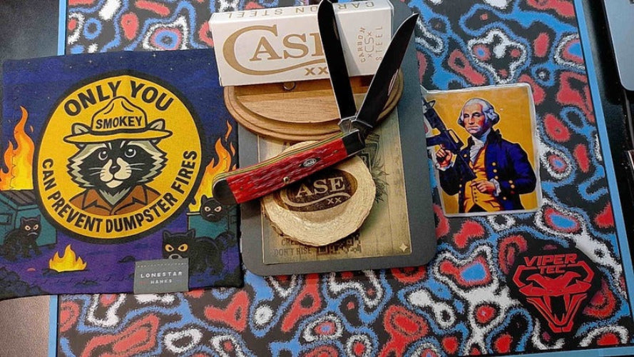 A flat-lay display of various Locomoter Games collectible items, including a 'Only You Smokey' sticker, a folding pocket knife with a red handle, a 'CASE' branded coaster, a 'Smoky' card, and a red 'Viper' logo patch, all arranged on a colorful patterned blanket.
